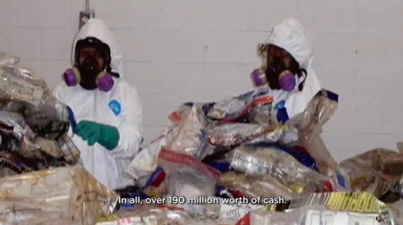 Two people in hazmat suits sort through bags of discolored currency