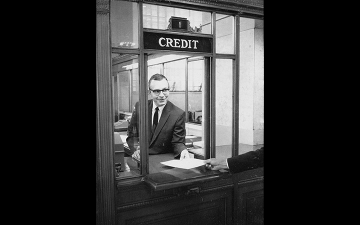 Bernie Berns hands paper through the Credit window on the first floor of the Federal Reserve Bank of St. Louis, 1967 (via <a href="https://fraser.stlouisfed.org/archival-collection/federal-reserve-bank-st-louis-centennial-5182/credit-discount-window-st-louis-fed-photograph-527398">FRASER</a>)
